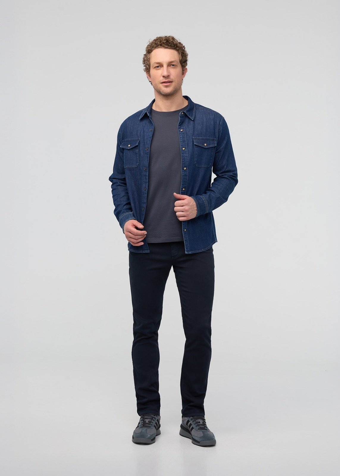 Man wearing a blue denim shirt over a gray t-shirt and black pants on a light gray background