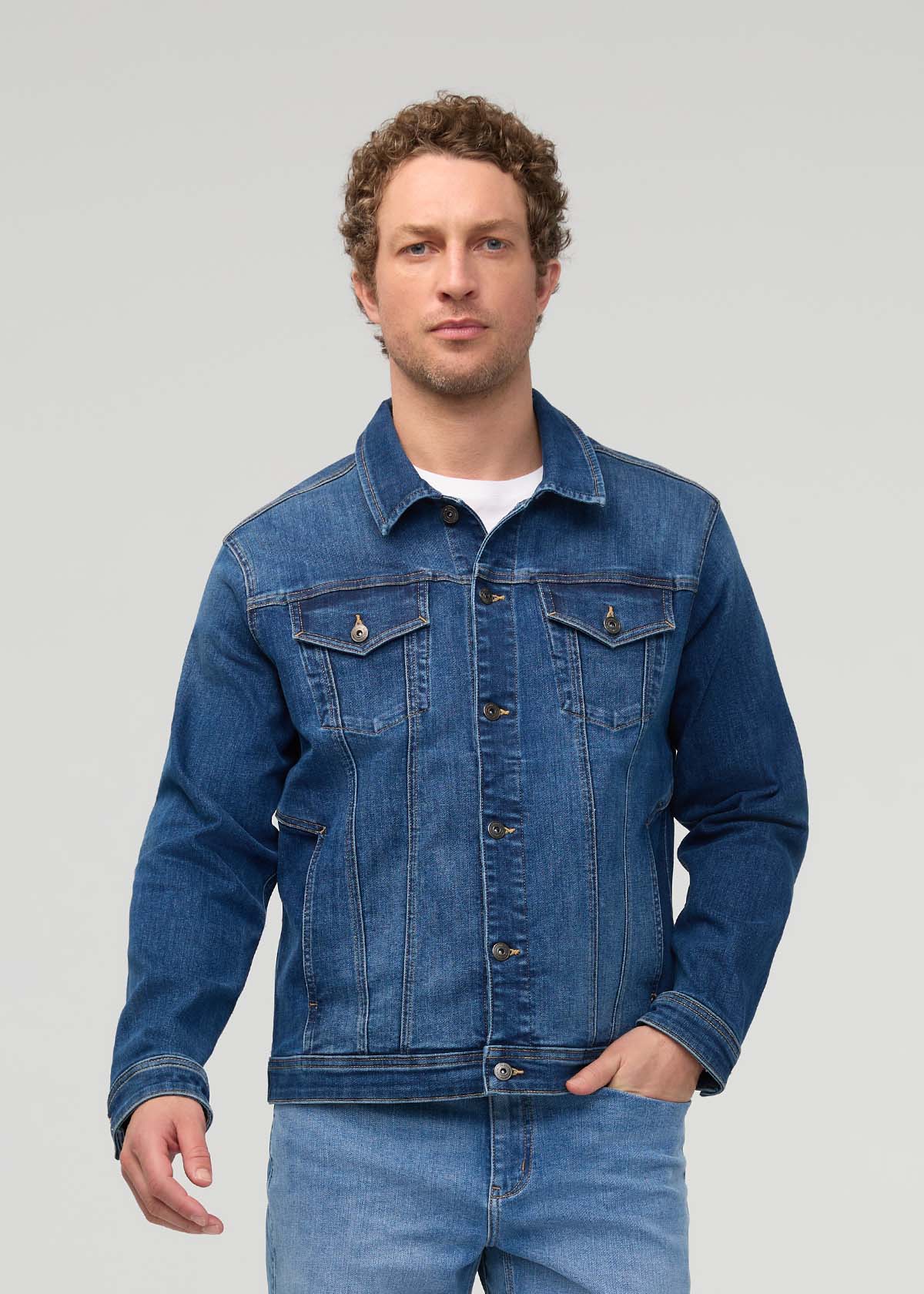 Man wearing dark indigo denim trucker jacket with button front, chest pockets, and rolled cuffs over white t-shirt.