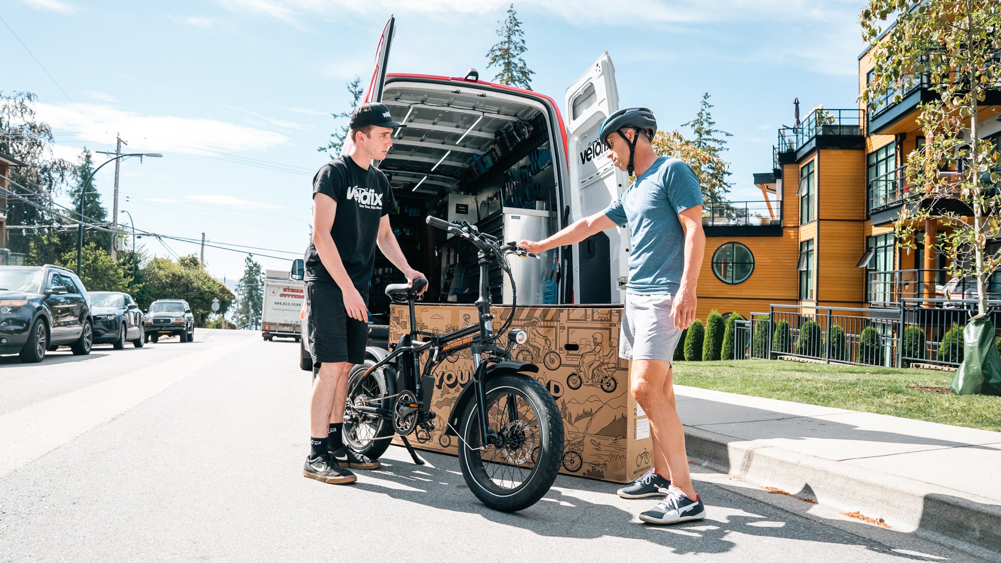 Person in black shirt and shorts stands by a van talking to a person in a light blue shirt, gray shorts, and a bike helmet, who is holding a black bike. Suburban street background.