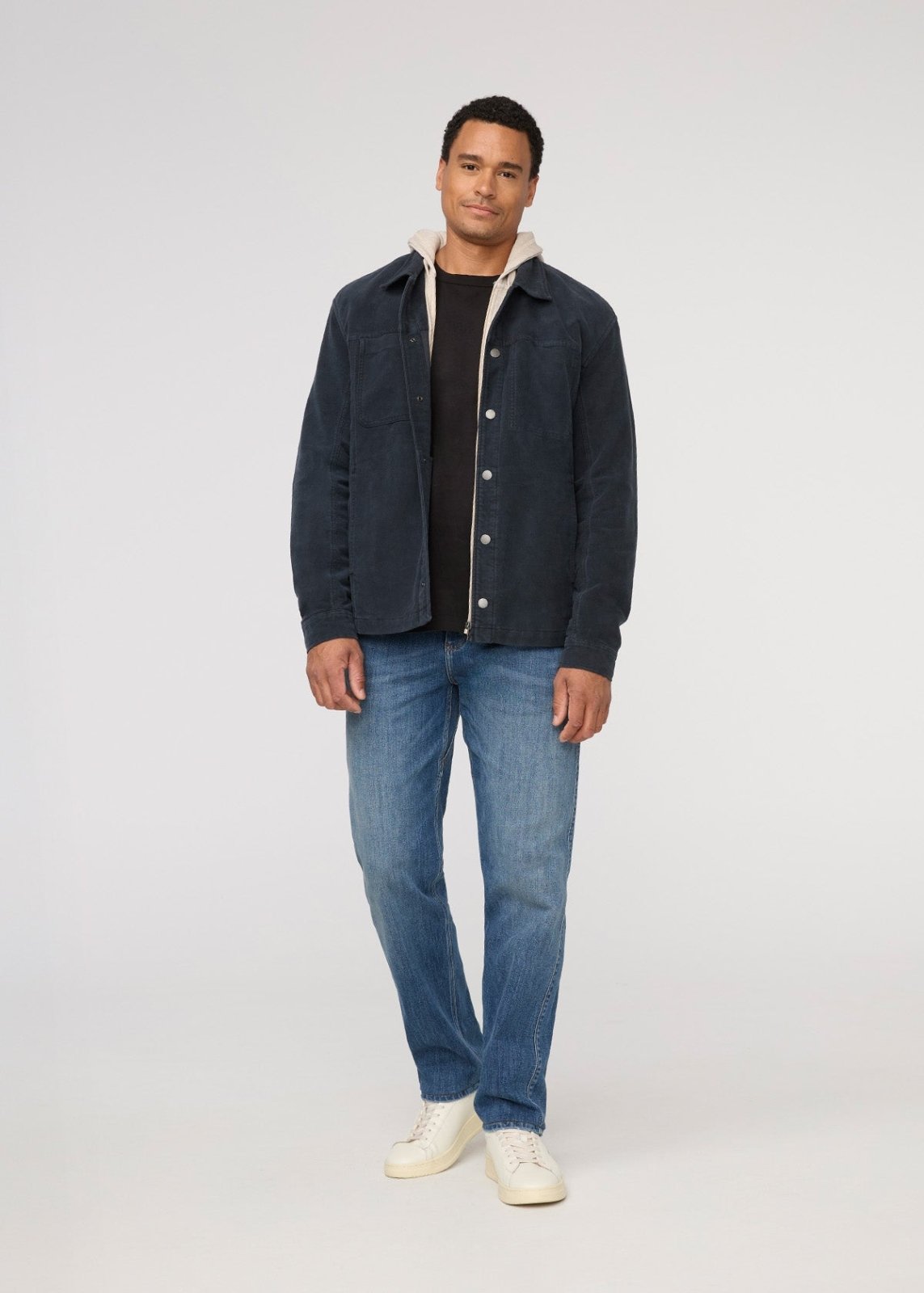 Man wearing a dark jacket, black shirt, blue jeans, and white shoes on a light gray background