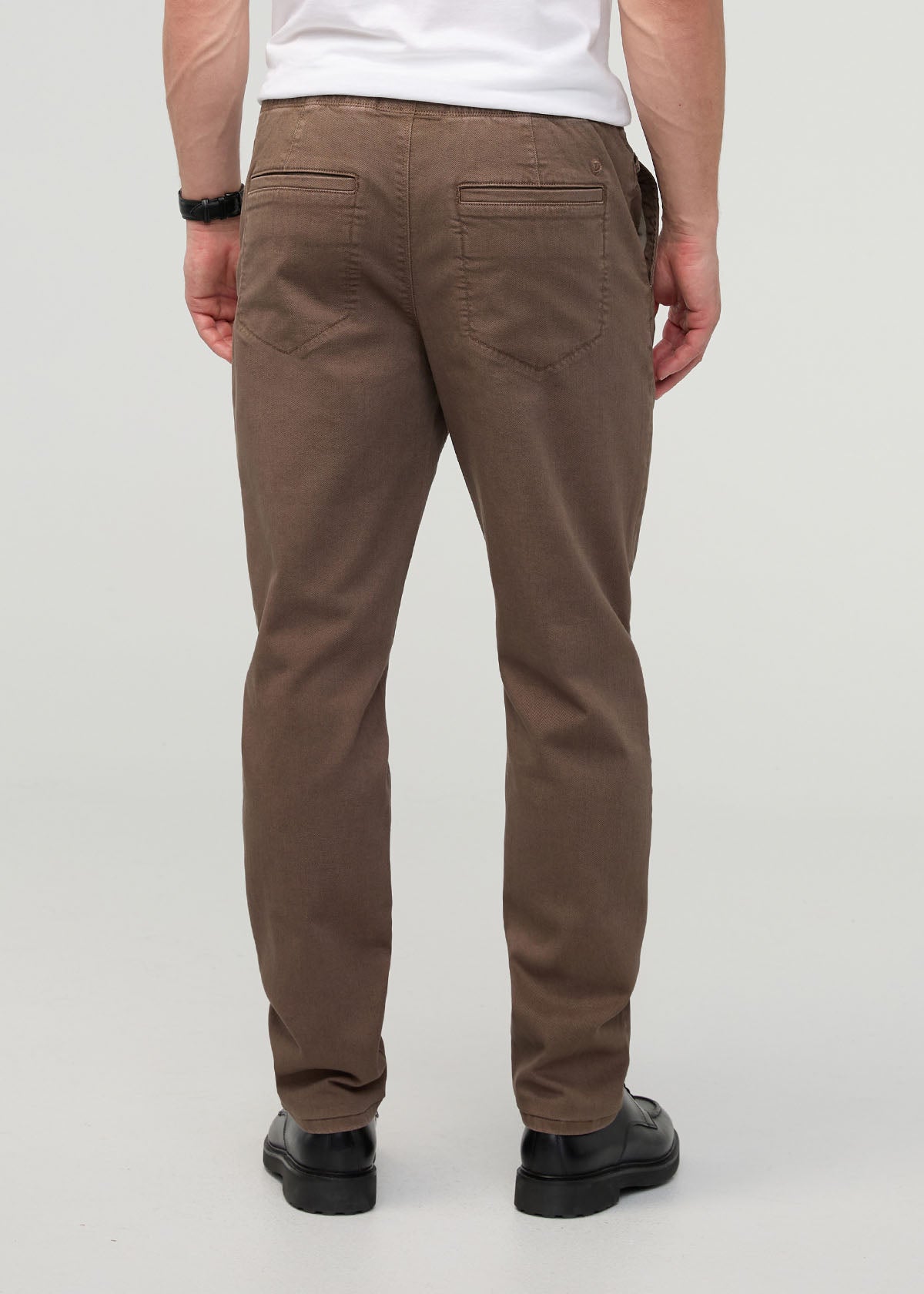 Man wearing brown relaxed pull-on pants with back pockets, white shirt, and black shoes, shown from behind.